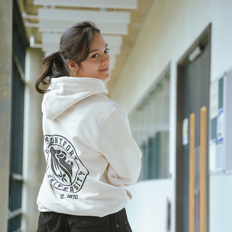A woman stands with her back to the camera and smiles forward, she wears a white hoodie with black DMU branding on the back