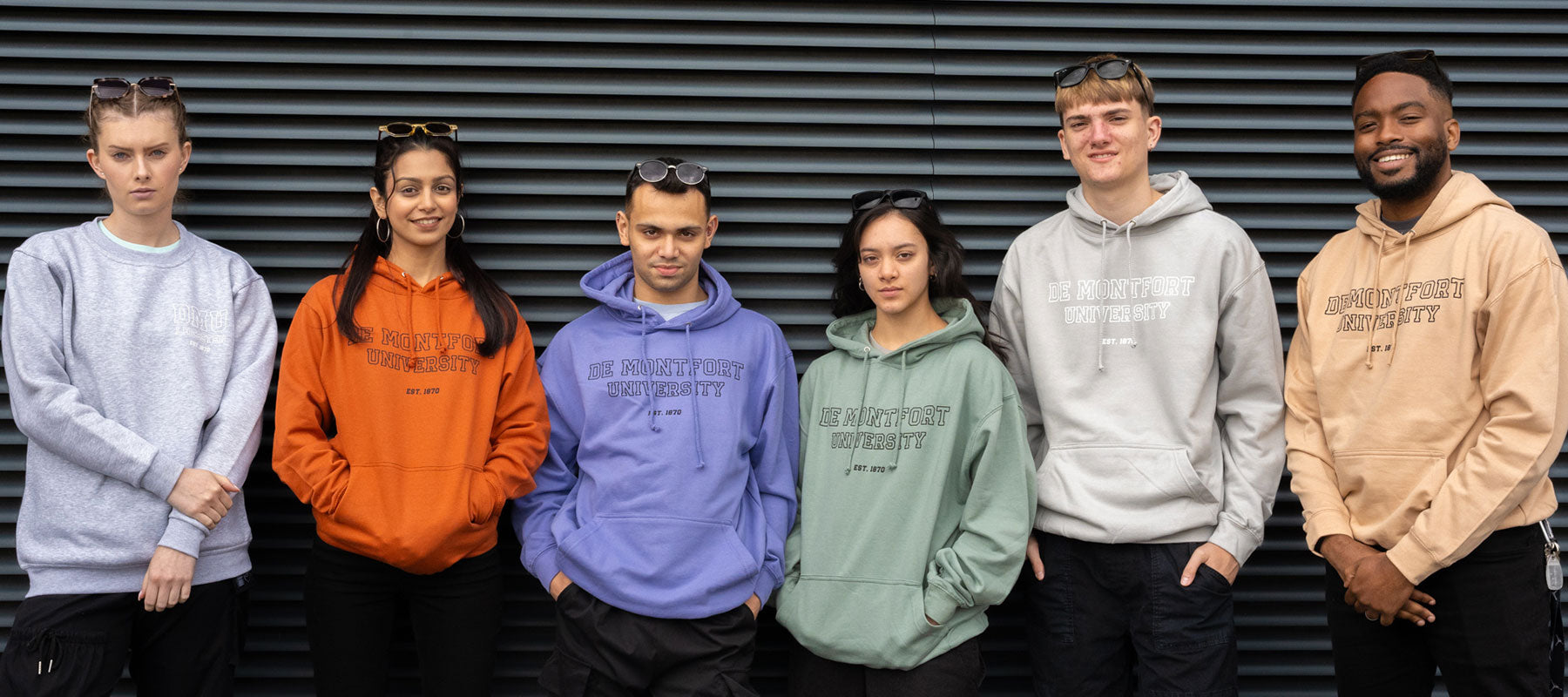 Six young people stand in front of grey shutters wearing a range of bright coloured branded hoodies and sweatshirts